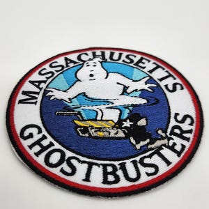 Peut inclure: &Eacute;cusson brod&eacute; circulaire avec le logo Ghostbusters. Le design comprend un fant&ocirc;me blanc, un fond bleu et les mots "Massachusetts Ghostbusters" en cercle. L'&eacute;cusson a une bordure rouge.