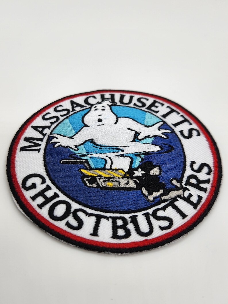 Peut inclure: &Eacute;cusson brod&eacute; circulaire avec le logo Ghostbusters. Le design comprend un fant&ocirc;me blanc, un fond bleu et les mots "Massachusetts Ghostbusters" en cercle. L'&eacute;cusson a une bordure rouge.