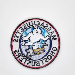 Peut inclure: &Eacute;cusson rond avec un fant&ocirc;me blanc sur fond bleu et blanc. Le patch est bord&eacute; d'un anneau rouge et noir, avec les mots "MASSACHUSETTS GHOSTBUSTERS" dispos&eacute;s en cercle autour du fant&ocirc;me.