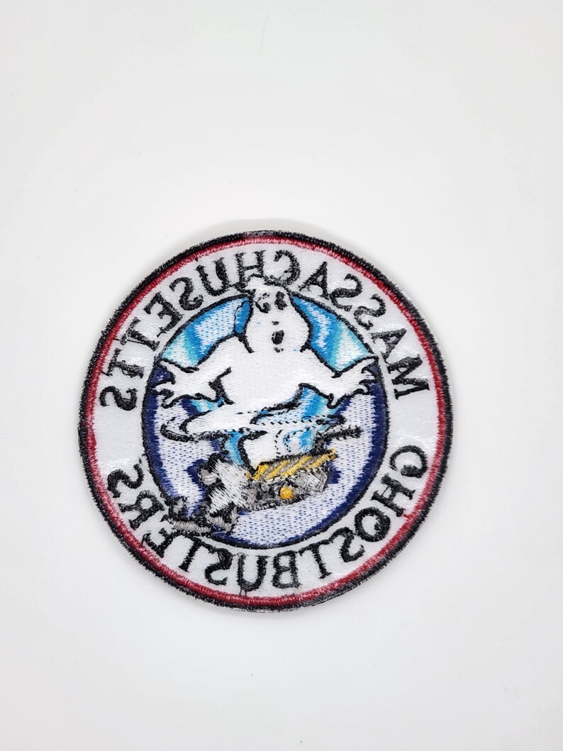Peut inclure: &Eacute;cusson rond avec un fant&ocirc;me blanc sur fond bleu et blanc. Le patch est bord&eacute; d'un anneau rouge et noir, avec les mots "MASSACHUSETTS GHOSTBUSTERS" dispos&eacute;s en cercle autour du fant&ocirc;me.