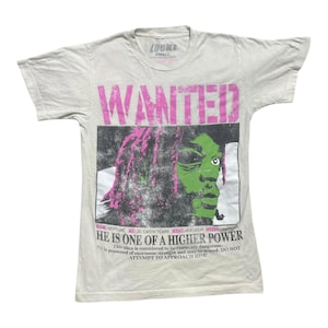 May include: Off-white t-shirt with a graphic print. The print features the word "WANTED" in pink above an image of a green-skinned figure with pink hair. Text below reads "HE IS ONE OF A HIGHER POWER".