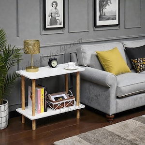 May include: A white side table with two shelves and wooden legs. A gold-coloured lamp, a black alarm clock, and a decorative basket are on the table. The table is next to a grey sofa with yellow and black cushions.