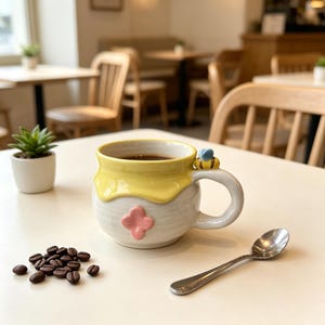 Op de afbeelding: Een keramische koffiemok met een gele honingachtige rand en een roze bloemmotief. Een kleine bij versiert het handvat. De mok is gevuld met koffie en staat op een witte tafel naast een zilveren lepel en koffiebonen.