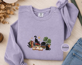 May include: A light purple crewneck sweatshirt featuring an embroidered graphic of three black cats, a stack of colorful books, a steaming cup of coffee, and a potted monstera plant, accented with small yellow stars on a soft fabric.