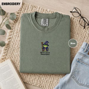 May include: A moss green crewneck t-shirt featuring an embroidered graphic of a small green frog wearing a blue wizard hat and robe, holding a wooden staff. Below the frog, the text reads Off to commit Shenanigans in black lettering.