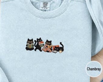 May include: A light blue crewneck sweatshirt featuring an embroidered graphic of four black cats reading books. The cats are depicted with glasses, holding stacks of books, sitting under a blanket, and using a tablet next to a small coffee mug.
