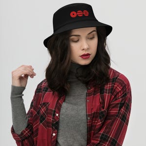 May include: Black bucket hat with a red circular logo. The hat is made of a soft material and has a wide brim. The hat is worn by a person wearing a red and black plaid shirt and a gray turtleneck.