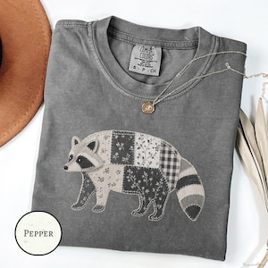 May include: A charcoal gray short-sleeve t-shirt featuring a large raccoon applique made of various fabric patches with floral and checkered patterns. The shirt has a crew neckline with a Comfort Colors label and a small circular pendant necklace.