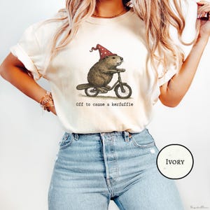 Puede incluir: Camiseta de color marfil con una ilustración caprichosa de un castor montando en bicicleta y con un sombrero rojo de lunares. El texto "Off to cause a kerfuffle" está impreso debajo. La camiseta es de material suave.