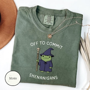 Może przedstawiać: Zielona koszulka w kolorze mchu z napisem "OFF TO COMMIT SHENANIGANS" i kreskówkową żabą w kapeluszu i szacie czarodzieja. Żaba trzyma laskę. Złoty naszyjnik jest zarzucony na koszulkę.