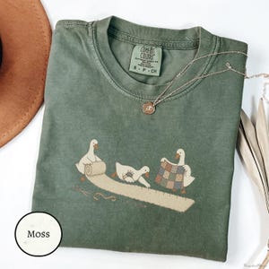 May include: A moss green short-sleeve t-shirt featuring a graphic of three white geese engaged in sewing activities, including unrolling fabric and holding a quilt. The shirt has a crew neckline and a Comfort Colors brand label at the inner collar.