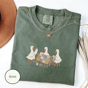 Puede incluir: Una camiseta de manga corta color verde musgo con un gráfico de tres gansos blancos sentados en taburetes de madera acolchando una manta de patchwork colorida. La camiseta tiene una etiqueta de la marca Comfort Colors en el cuello.