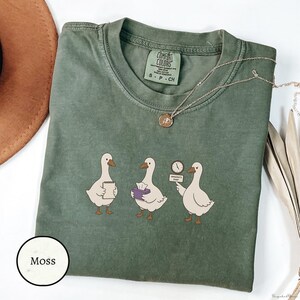 Puede incluir: Una camiseta de cuello redondo color verde musgo con un gráfico de tres gansos blancos. El primer ganso sostiene un bloc de notas, el segundo una caja de pañuelos y el tercero un reloj con un cartel que dice session's over. Etiqueta Comfort Colors.