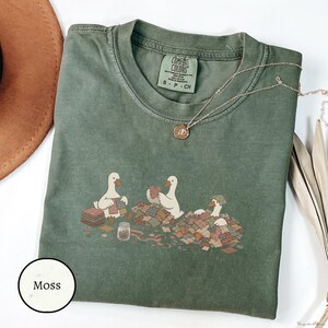 May include: A moss green short-sleeved t-shirt featuring a graphic of three white geese sewing with colourful fabric scraps. The shirt has a crew neckline and a Comfort Colors label at the inner collar. A delicate gold-toned necklace rests on the fabric.