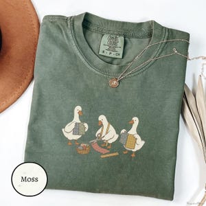 May include: A moss green short-sleeve t-shirt featuring a graphic of three white geese engaged in sewing activities, including holding fabric, using scissors, and a basket of scraps. The shirt has a Comfort Colors label at the inner neckline.