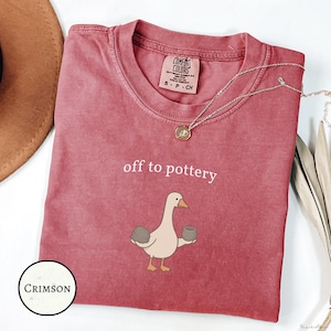 Op de afbeelding: Een karmozijnrood t-shirt met korte mouwen en een ronde hals, voorzien van de tekst "off to pottery" in witte letters boven een afbeelding van een gans die een grijze klomp klei en een kleine grijze pot vasthoudt. Het shirt heeft een Comfort Colors label.