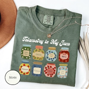 May include: Moss green Comfort Colors t-shirt with the text "Telemetry is My Jam." The shirt features illustrations of eight jam jars, each labeled with medical terms like "Rhythm Watch" and "Alerts." A silver necklace with a pendant is draped on the shirt.