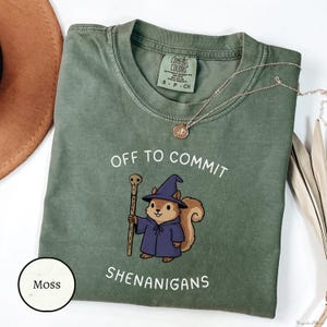 May include: Moss green t-shirt with the text "OFF TO COMMIT SHENANIGANS" and a cartoon squirrel wearing a wizard hat and robe. The shirt has a crew neck and short sleeves. A gold necklace is draped across the shirt.