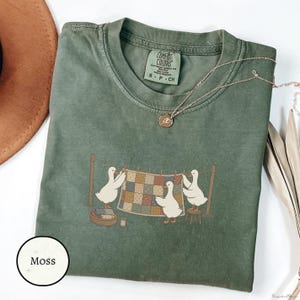 Puede incluir: Una camiseta de manga corta color verde musgo con cuello redondo y etiqueta de la marca Comfort Colors. El gráfico muestra tres patos blancos colgando una colcha de patchwork multicolor en un tendedero, con una cesta y un taburete.