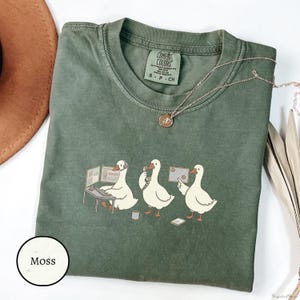 May include: A moss green crewneck t-shirt featuring a graphic of three white geese in office settings. One goose wears a headset at a computer, one holds a telephone receiver, and one points at a document. The shirt has a Comfort Colors brand label.