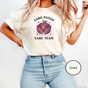 May include: Ivory-coloured t-shirt with a graphic of a heart inside a cabbage, and the text "CABG PATCH CARE TEAM". The design includes a heartbeat line and medical symbols. The shirt is a crew neck.