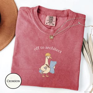 May include: A crimson t-shirt featuring a graphic of a goose wearing a yellow hard hat, holding a hammer and blue architectural blueprints. The text above the goose reads off to architect. The shirt has a Comfort Colors brand neck label.