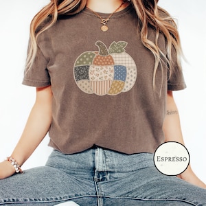 Può includere: Una t-shirt a maniche corte marrone scuro con una grande grafica centrale raffigurante una zucca composta da vari motivi di tessuto in stile patchwork in tonalità tenui di verde, arancione, blu e crema, con la parola ESPRESSO in un cerchio.