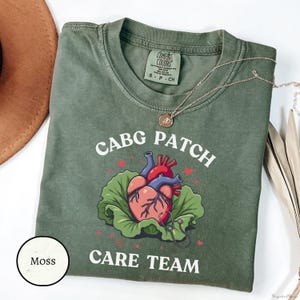 May include: Moss green t-shirt with the words "CABG PATCH CARE TEAM" and a graphic of a heart on a bed of lettuce. A gold necklace is draped across the shirt. The shirt is labelled "Comfort Colors" and size S-P-CH.