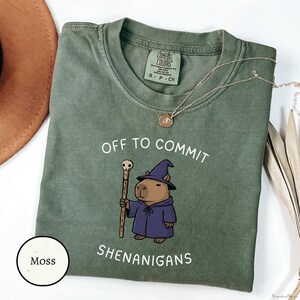 Puede incluir: Camiseta verde musgo con el texto "OFF TO COMMIT SHENANIGANS" y un dibujo de un carpincho con un atuendo de mago, sosteniendo un bastón. Un collar dorado está sobre la camiseta.