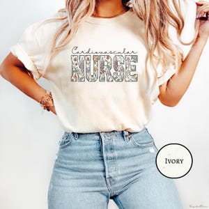 May include: Ivory-coloured t-shirt featuring the words "Cardiovascular Nurse" in a floral design. The text is in a block letter style, with "Nurse" being the largest. The shirt is a crew neck style.