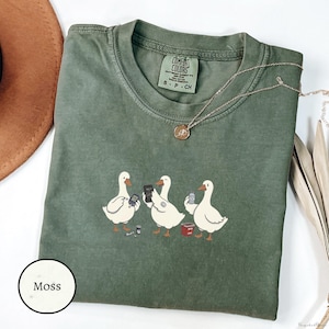 Könnte beinhalten: Ein moosgrünes Comfort Colors T-Shirt mit einer Grafik von drei weißen Enten, die jeweils einen Gegenstand halten. Eine Ente hält ein Telefon, eine andere ein Getränk und die dritte ein Buch. Eine goldene Halskette liegt über dem Shirt.