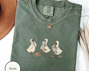 May include: A moss green crewneck t-shirt featuring a graphic of three white ducks. One duck eats bread, another examines a box with a magnifying glass, and the third holds a box labelled Gluten Free Crackers. The shirt has a Comfort Colors label.