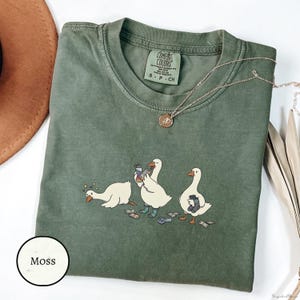 May include: A moss green short-sleeved t-shirt featuring a graphic of three white geese. One goose is lying down with stars above its head, one is holding a water bottle, and one is holding a pair of socks. The shirt has a Comfort Colors brand label.
