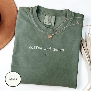 May include: A moss green t-shirt with the words "coffee and jesus" and a cross graphic in white. A gold necklace with a pendant is draped over the shirt. The shirt is folded on a white surface.
