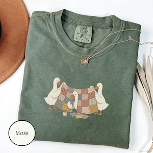 Puede incluir: Una camiseta de manga corta color verde musgo con cuello redondo y etiqueta Comfort Colors. El gráfico muestra tres patos blancos sosteniendo una colcha de retazos multicolor, con un carrete de hilo y retazos de tela en el suelo.