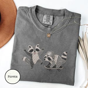 May include: A charcoal gray crew neck t-shirt featuring a graphic of two raccoons. One raccoon is jumping, while the other wears a colander on its head and rides a skateboard. The shirt has a Comfort Colors label and a small gold-toned necklace.