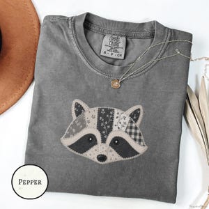 May include: A charcoal gray short-sleeve t-shirt featuring a central graphic of a raccoon face composed of various patchwork patterns including floral and gingham prints. The shirt has a crew neckline and a Comfort Colors brand label inside.