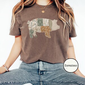 Puede incluir: Una camiseta de manga corta color marrón oscuro con un gráfico de un cerdo compuesto por varios patrones de patchwork en tonos verde salvia, crema y bronceado. El diseño del cerdo incluye texturas florales, de cuadros y sólidas. Una etiqueta dice ESPRESSO.