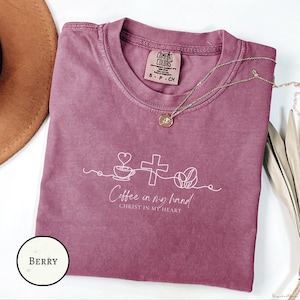 Op de afbeelding: Bessenkleurig T-shirt met de tekst "Coffee in my hand, Christ in my heart" in witte letters. Het shirt heeft een lijntekening van een koffiekopje, een kruis en een koffieboon. Een gouden ketting is over het shirt gedrapeerd.