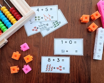 Puede incluir: Un ábaco de madera con cuentas de colores, varias tarjetas de matemáticas plastificadas con sumas e ilustraciones de animales, y varios cubos de plástico encajables en colores naranja, rosa y blanco para practicar aritmética.