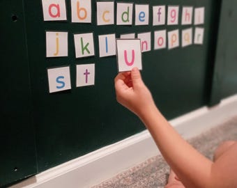Puede incluir: Un tablero verde oscuro con filas de pequeñas tarjetas cuadradas blancas con letras minúsculas coloridas del alfabeto. Una mano sostiene una tarjeta cuadrada blanca con la letra mayúscula U en rosa, mientras otras letras están en el suelo.