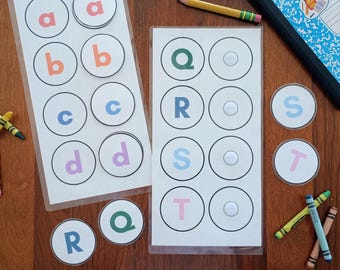 Puede incluir: Dos hojas laminadas con filas de recortes circulares que contienen letras minúsculas y mayúsculas en varios colores, incluyendo rojo, naranja, azul, morado, verde y rosa. Varias fichas circulares con letras están colocadas cerca.