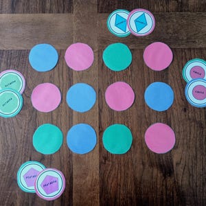 Pode incluir: Uma coleção de fichas educativas circulares em azul, rosa e verde. Algumas fichas apresentam formas geométricas e etiquetas incluindo losango, octógono, círculo e pentágono. As fichas estão dispostas em grade sobre uma superfície de madeira.