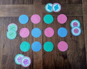 Puede incluir: Una colección de fichas educativas circulares en azul, rosa y verde. Algunas fichas presentan formas geométricas y etiquetas que incluyen rombo, octágono, círculo y pentágono. Las fichas están dispuestas en un patrón de cuadrícula sobre madera.