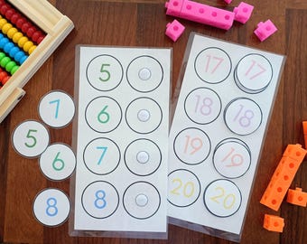 Puede incluir: Set de aprendizaje matemático que incluye dos hojas laminadas con tarjetas circulares numeradas del 5 al 20, un ábaco de madera con cuentas de colores y varios cubos de plástico entrelazables en rosa y naranja para contar.