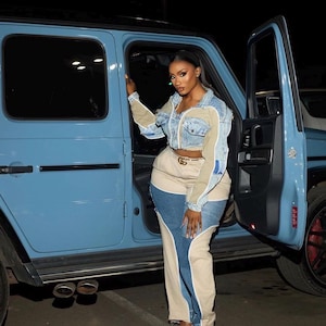 May include: A woman in a denim jacket with beige and blue panels, and colour-block jeans. She wears a gold belt and black sandals. A light blue SUV is visible in the background.