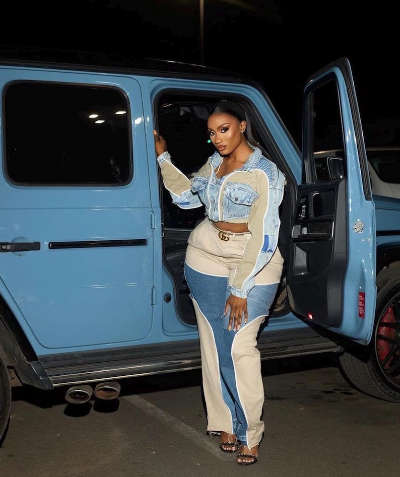 May include: A woman in a denim jacket with beige and blue panels, and colour-block jeans. She wears a gold belt and black sandals. A light blue SUV is visible in the background.