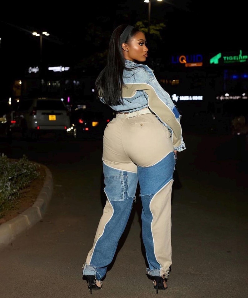 May include: A woman in a denim jacket with beige and blue panels, and patchwork jeans in beige and blue. She is wearing black high-heeled shoes. The background shows city lights at night.