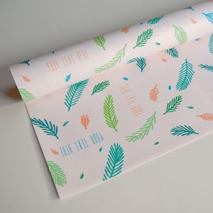 May include: A roll of light pink wrapping paper featuring a repeating pattern of stylised fern leaves in shades of teal, lime green, and orange. The paper includes the repeated text "YOUR TEXT HERE" printed in a simple, sans-serif font.