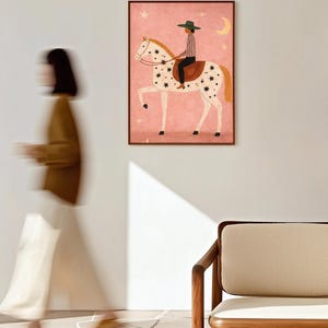 May include: A framed art print depicts a person riding a white horse adorned with black star-shaped spots. The rider wears a green hat and striped shirt. The background is pink with stars and a crescent moon.